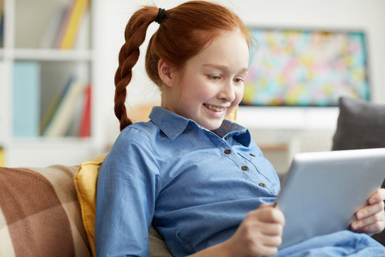 Portrait of smiling teenage girl using digital tablet while lying on sofa, watching videos or browsing internet, copy space