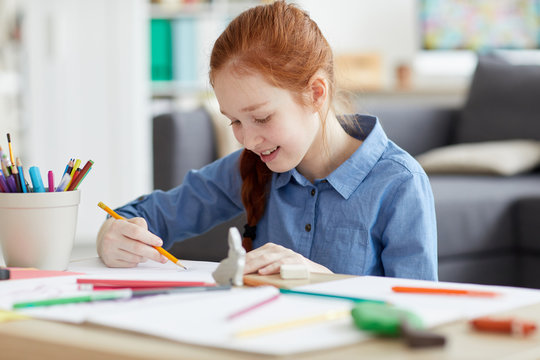 Portrait of red haired teenage girl drawing pictures sitting at table at home and smiling, copy space