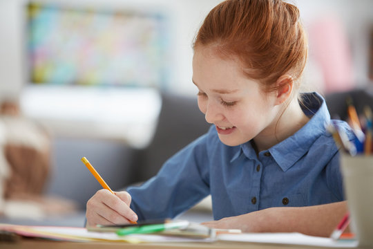 Portrait of red-haired schoolgirl studying sitting at desk and writing with pencil, copy space