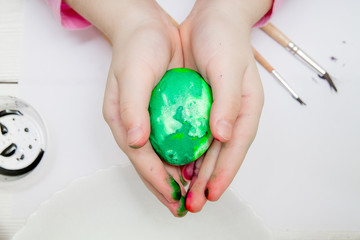 A flat lay with kid's hands painting Easter eggs on white wooden table