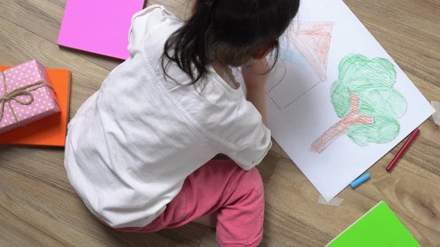 The Asian little girl is drawing the picture in the paper on the wooden ground she has many book and color pencil around her, top view of child on floor, Educational concept for school kids