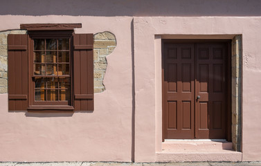 Brown wood framed window with brown shutters set against a pink wall with a brown wooden door beside it with some stone wall 
