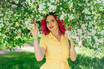 Beautiful and young woman with bright red hair and ref lips standing next to a blooming apple tree...