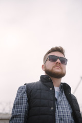 A man with a beard and sunglasses in a plaid shirt and waistcoat posing on the street to advertise men's clothing. Advertising menswear