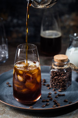 Ice coffee cold summer drink in a tall glass with pouring coffee and coffee beans on a stone background.
