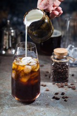 Ice coffee cold summer drink in a tall glass with pouring cream and coffee beans on a stone background.