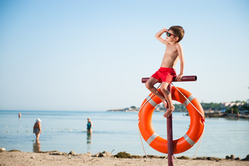 Fototapeta na wymiar beautiful small kid in sunglasses sitting on pole with orange lifebuoy hanging on it on summer beach baywatch