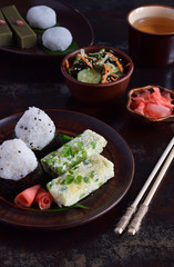 Mix of Japanese food - rice balls onigiri, omelette, ginger, sunomono wakame cucumber salad. Traditional dessert of bean and green tea matcha - jelly yokan, daifuku mochi. Asian breakfast or lunch