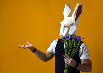 man in paper rabbit mask holds a bouquet of irises flowers on a yellow studio background.