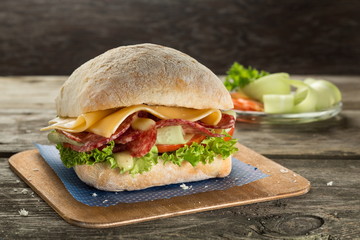 Ciabatta sandwich with salami, cheese, tomato and green salad close up on the wooden background