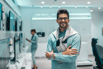 Fototapeta na wymiar Smiling mixed race man dressed elegant standing in tech store and holding arms crossed.