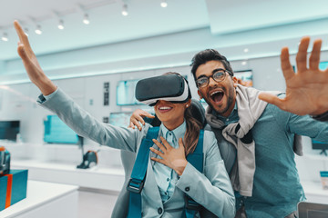Smiling Caucasian woman trying out virtual reality technology while her husband hugging her and looking at camera.