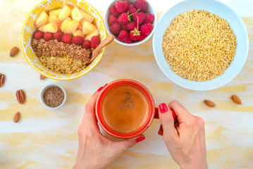 Top view, female hands holding a cup of coffee, a spoon with oatmeal, with honey nuts, raspberries, on a yellow wooden table Good morning - healthy breakfast background