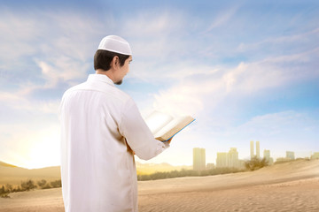 Rear view of asian muslim man with cap standing and reading the quran on the sand