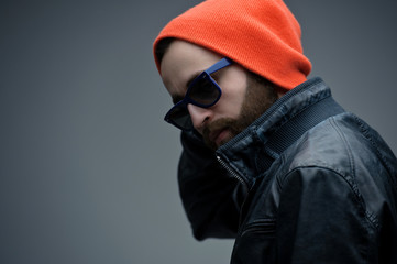 Profile portrait of a bearded brutal man in sunglasses and orange hat and black leather jacket, isolated on a dark grey background. Confident style.