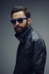 Profile portrait of a attractive bearded brutal man in sunglasses and leather jacket, isolated on a dark grey background. Confident style.