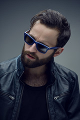 Profile portrait of a bearded brutal man in sunglasses and leather jacket, isolated on a dark grey background. Confident style.
