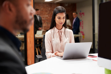 Businesswoman Working At Desk On Laptop In Open Plan Office With Colleagues In Background
