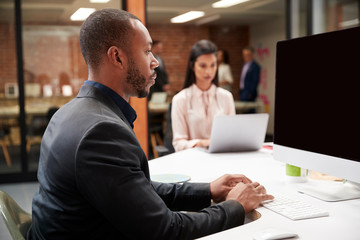 Businessman Working At Desk On Computer In Open Plan Office With Colleagues In Background