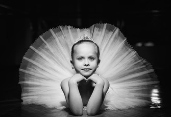 Czarno-białe zdjęcie uroczej uśmiechniętej baleriny w białej spódniczce baletnicy i koronie leżącej na podłodze z rękami pod brodą. Piękna mała dziewczynka kłaść w ładnej spódniczce baletnicy na czarnym tle. - 257404026