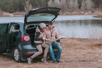 Young romantic couple is sitting in open trunk of car near the river and watching road map.