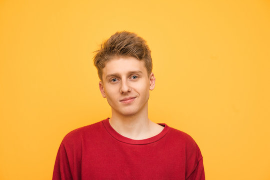 Handsome young man in a red sweatshirt stands on a yellow background, looks at the camera and smiles. Portrait of a happy teenager on a yellow background.