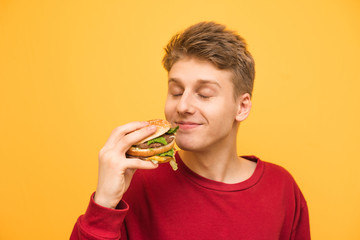Satisfied guy with his eyes closed and with a burger in his hands on a yellow background. Young man...