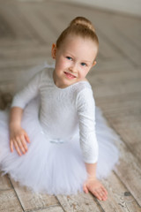 Fototapeta premium A small sweet ballerina in a white tutu and a body sitting on the birchwood floor in a light studio. Studio. Children and ballet