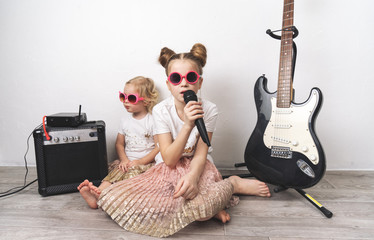 Two girls in pink sunglasses and identical T-shirts sing with a microphone and play the electric...