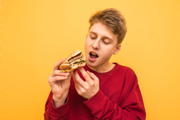 Portrait of a hungry guy looking at an appetizing buggy burger in his hand and going to eat. A very hungry young man eats a burger on a yellow background, isolated. Copyspace