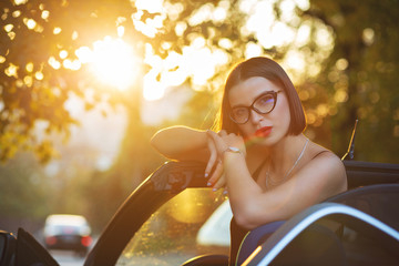 Gorgeous model wearing glasses, sitting in a convertible car with gold evening sunlight. Space for...