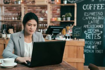 Fototapeta na wymiar Young asian businesswoman wearing grey suit using laptop computer sitting in cafe bar. Female working on netbook on wooden table with cup of hot coffee waiting for customer. lady having meeting.