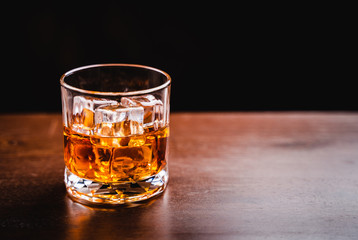 Glass of whiskey with ice cube on wooden table over black background