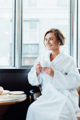 attractive brunette woman in bathrobe sitting and holding cup of coffee