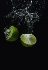 Fototapeta na wymiar Lime green fruit that is falling and has water spreading with a black background.