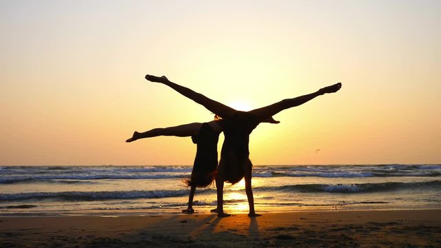 Silhouette of sporty young women practicing acrobatic on the beach at sunset. Mom and daughter doing gymnastic exercises synchronously together. Side view.