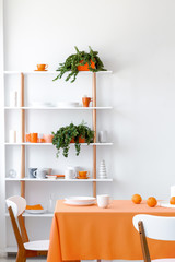 Wooden rack with candles, mugs and fresh plants in real photo of white dining room interior with table with orange cloth, cup and plates