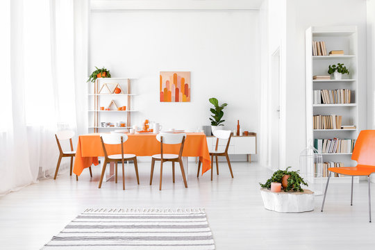 White flat interior with window with curtains, bookcase, rack with decor and long dining table with orange cloth and four chairs in the real photo