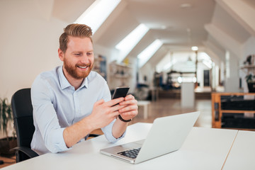 Positive ginger businessman using smartphone while sitting at the desk with laptop.