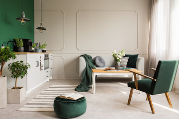 Open book on dark green velvet pouf in grey living room and kitchen interior with retro armchair,...