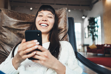 Portrait of handsome asian girl sitting in bright spacious room with mobile phone in hands