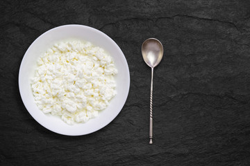 An overhead photo of fresh natural cottage cheese in a white ceramic bowl on the black stone desk. Old vintage silver spoon. Organic eco healthy meal, dairy product. Top view, photo from above.