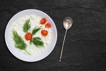 An overhead photo of fresh natural cottage cheese in a white ceramic bowl on the black stone desk. Green dill, vintage silver spoon and red tomatoes. Organic eco healthy meal, dairy product. Top view.
