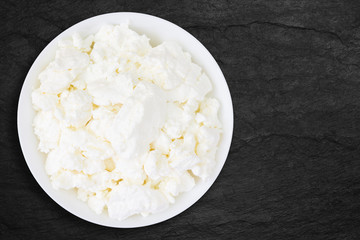 An overhead photo of fresh natural cottage cheese in a white ceramic bowl on the black stone desk. Organic eco healthy meal, dairy product. Top view. Place for text, copyspase.