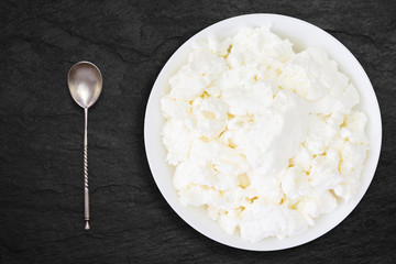 An overhead photo of fresh natural cottage cheese with the vintage old silver spoon in a white ceramic bowl on the black stone desk. Organic eco healthy meal, dairy product. Top view.