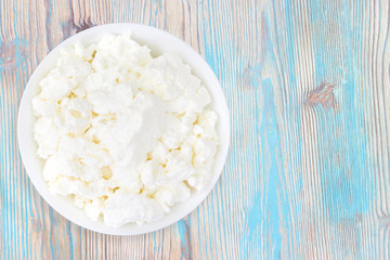 An overhead photo of fresh natural cottage cheese in a white ceramic bowl on the wooden table. Organic eco healthy meal, dairy product. Top view. Place for text, copyspace.