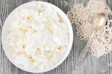 Fresh natural cottage cheese in a white ceramic bowl on the green wooden table. Organic eco healthy meal, dairy product. Top view, overhead photo. Rustic background with crochet doily and old spoon.