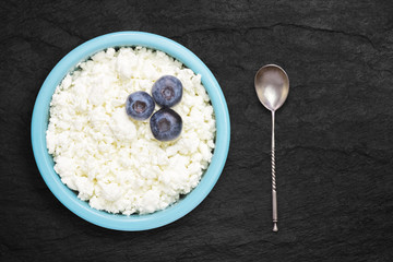 An overhead photo of fresh natural cottage cheese with blueberries and a silver spoon in a blue ceramic bowl on the black stone desk. Organic eco healthy meal, dairy product. Top view with copyspase.