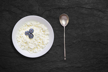 Organic eco healthy meal, dairy product. An overhead photo of fresh natural cottage cheese with blueberries and a silver spoon in a white ceramic bowl on the black stone desk. Top view with copyspase.
