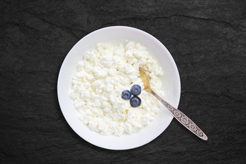 Organic eco healthy meal, dairy product. An overhead photo of fresh natural cottage cheese with blueberries and a silver spoon in a white ceramic bowl on the black stone desk. Top view with copyspase.